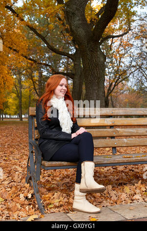 Redhead girl in autunno city park, sedersi sul banco di legno, uno persone al giorno, foglie di Giallo autunno Foto Stock