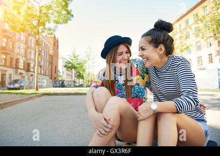 Due piuttosto alla moda giovane donna seduta ridendo e scherzando su uno skateboard in una tranquilla strada urbana con spazio di copia Foto Stock