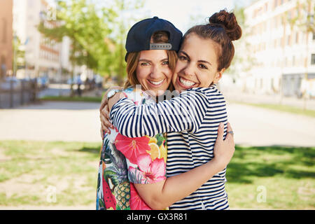 Due felici piuttosto giovani amici di sesso femminile con vivaci sorrisi che abbraccia ogni altra in un abbraccio in un tranquillo contesto urbano Foto Stock