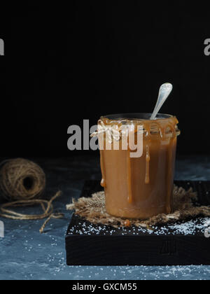 In casa salato salsa di caramello in un rustico il vasetto di vetro, cosparso sale. Sfondo nero Foto Stock