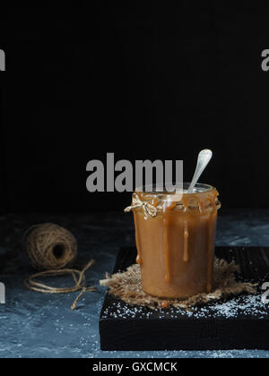 In casa salato salsa di caramello in un rustico il vasetto di vetro, cosparso sale. Sfondo nero Foto Stock