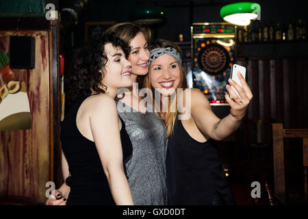 Tre femmina adulta amici tenendo lo smartphone su selfie notte fuori in bar Foto Stock