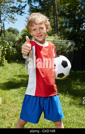 Ritratto di ragazzo indossa uniformi di calcio tenendo palla calcio e dando i pollici in su in giardino Foto Stock