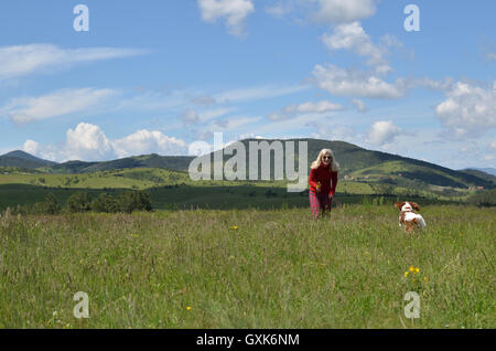 Gioiosa donna bionda in rosso che esercitano con il suo cane (Cavalier King Charles Spaniel) su un prato e il meraviglioso paesaggio Foto Stock