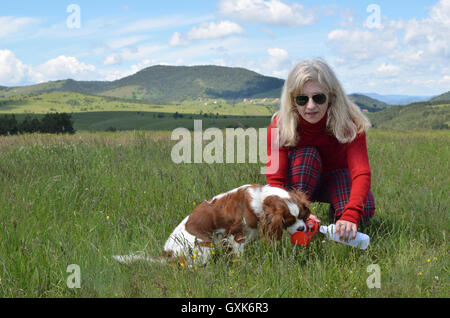 Donna con occhiali da sole sta aiutando il suo cane di bere acqua dal portatile erogatore di acqua in un prato di montagna Foto Stock