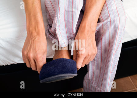 Primo piano di un giovane uomo in pigiama seduto sul bordo del letto a mettere su un paio di pantofole caldi Foto Stock