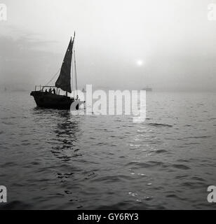 Anni '50, storico, notturno e una spazzatura (barca) è ormeggiata in acqua al porto di Aberdeen, Hong Kong Island. Le giunche sono barche o navi orientali da pesca dove gli occupanti vivrebbero spesso su di loro. Il porto di Aberdeen è un noto "villaggio galleggiante", sede di molti fondali di barche spazzatura. Foto Stock