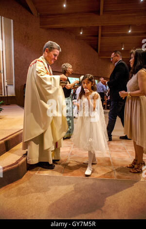 Un formalmente vestiti ragazza riceve la Comunione durante la Prima Comunione messa in una Laguna Niguel, CA, chiesa cattolica. La prima comunione è una cerimonia in alcune tradizioni cristiane durante il quale una persona riceve prima l Eucaristia. Nota derubato monsignor. Foto Stock