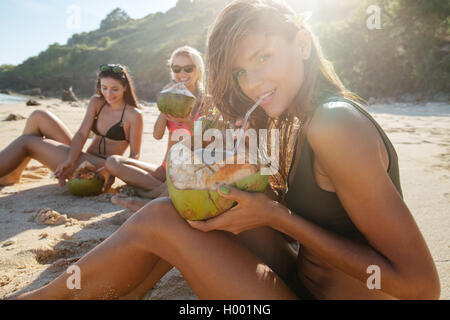 Ritratto di giovane e bella donna fresca potabile Acqua di cocco con i suoi amici seduti in background sulla spiaggia. Gruppo di voi Foto Stock