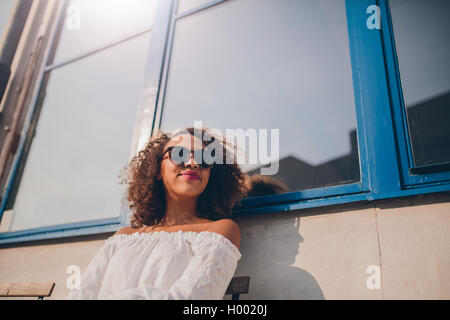 Colpo di attraente giovane donna africana seduti all'aperto e guardando lontano. Femmina felice indossando occhiali da sole seduto fuori. Foto Stock