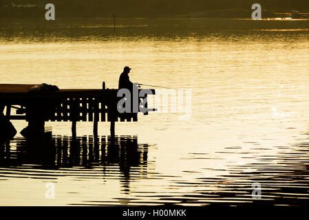 Silhouette di uomo seduto sul molo di sera la luce del sole durante la pesca. Foto Stock