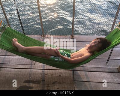 9 anni Eurasian ragazzo disteso su un verde amaca sulla piattaforma di legno vicino al mare di Hong Kong Foto Stock