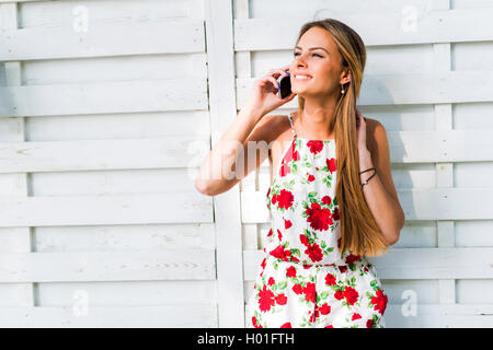 Bella giovane donna felice sorridente durante una chiamata telefonica mentre appoggiata contro un muro bianco Foto Stock