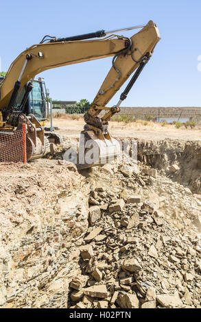 Escavatore giallo digger su un sito di costruzione scava attraverso pesanti pietre per creare uno spazio per la cantina e le fondamenta di una casa Foto Stock