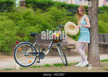 Attraente bionda womanwith cappello di paglia in piedi nel parco e posa vicino alla moto con cesto pieno di negozi di generi alimentari. Foto Stock