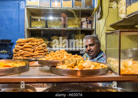 JODHPUR, India - 16 febbraio 2015: il fornitore si siede in negozio con vari alimenti su piastre metalliche e tagliatelle sui ripiani. Foto Stock