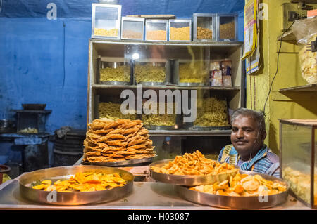 JODHPUR, India - 16 febbraio 2015: il fornitore si siede in negozio con vari alimenti su piastre metalliche e tagliatelle sui ripiani. Foto Stock