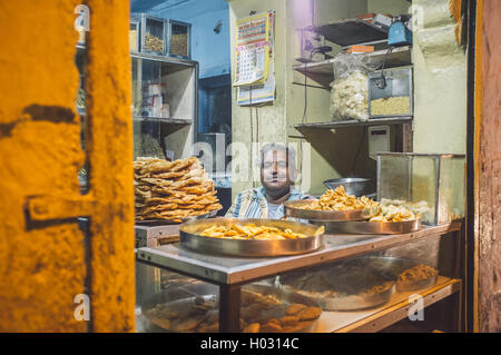 JODHPUR, India - 16 febbraio 2015: il fornitore si siede in negozio con gli alimenti sulle piastre in metallo. Post-elaborati con grano e texture. Foto Stock