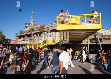 Una folla di gente che per godersi il sole al di fuori del Royal Festival Hall sulla sponda sud del Tamigi a Londra. Foto Stock