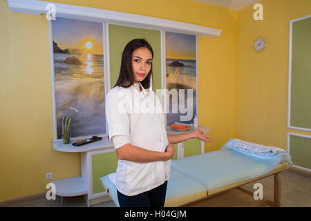 Sorridente terapista di bellezza in piedi accanto al lettino da massaggio presso il centro termale e accoglie invita il cliente Foto Stock