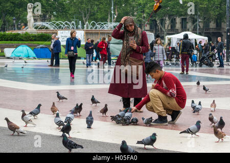 Ragazzo musulmano alimentare street piccioni in Barcellona, mentre il hijab-indossando la madre prende una foto di lui con il suo iPhone. Foto Stock