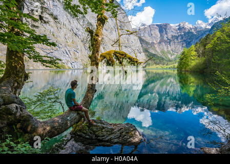 Giovane uomo seduto su albero sopra l'acqua, cercando in distanza, riflesso nel lago Obersee, Salet am Königssee Foto Stock