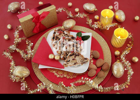 Natale ancora in vita con lo Stollen torta, confezione regalo, candele, agrifoglio , foil cioccolatini incartati e oro ninnolo decorazioni su rosso. Foto Stock