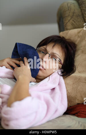 La mezza età stanco donna anestetico dopo tirando i denti di saggezza si raffredda il tuo volto, rilassante e giacente a casa senza trucco in rosa Foto Stock