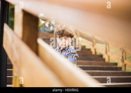 Ritratto di poco carino bambino seduto sulle scale di legno nel parco della città. Ragazzo guarda la telecamera attraverso parapetto in legno di scale Foto Stock