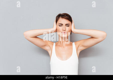 Giovane donna con gli occhi chiusi che copre le sue orecchie con palme isolate su uno sfondo grigio Foto Stock