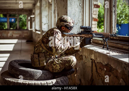 Uomo militare sniper con il fucile automatico con un mirino telescopico seduta dalla finestra nella costruzione Foto Stock