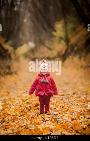 Bambina in rosso cappotto presso Scenic caduta park. Il bambino che indossa il cappuccio di maglia su sfondo del fogliame. Bambino bambino che gioca con il giallo Foto Stock