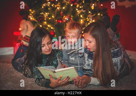 Famiglia giovane e il bambino vicino a un albero di natale in camera decorata in leggere il libro. Foto Stock