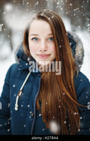 Giovane donna in abiti invernali in piedi sotto la neve, sorridente, guardando la fotocamera. In inverno il paesaggio della foresta e la caduta di neve sul b Foto Stock