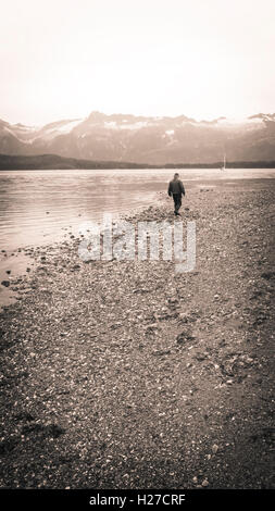 Uomo che cammina su una spiaggia nel sud-est dell Alaska con una barca a vela ancorata in background in modalità monocromatica con una vignette per un vintag Foto Stock