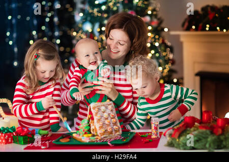 La famiglia felice, giovane madre con bambino, il piccolo ragazzo e ragazza rendendo gingerbread house al caminetto vicino albero di Natale Foto Stock