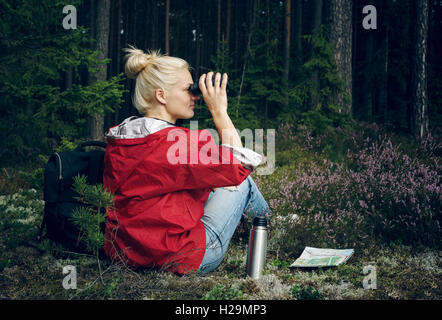 Giovane donna attiva tourist sedersi in una radura nel bosco con uno zaino, tenendo il binocolo e una mappa e guardando la fores Foto Stock