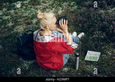 Giovane donna attiva tourist sedersi in una radura nel bosco con uno zaino, tenendo il binocolo e una mappa e guardando la fores Foto Stock