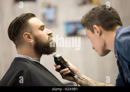 Vista di profilo ritratto del bel giovane barbuto uomo caucasico getting barba toelettatura in moderno barbiere. Parrucchiere con tatt Foto Stock