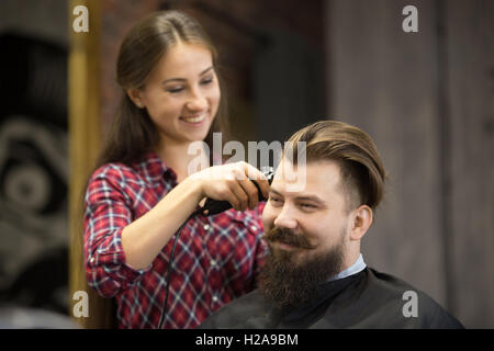 Ritratto di bello sorridente giovane barbuto uomo caucasico ottenere taglio di capelli alla moda nella moderna barbiere. Attraente ragazza barbiere wor Foto Stock