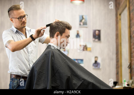 Shot interni del processo di lavoro in moderno barbiere. Vista laterale verticale del bel giovane ottenere taglio di capelli alla moda. Cool mal Foto Stock