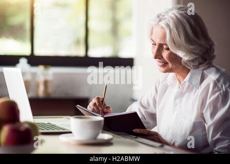 Grey-haired lady prendendo appunti con matita e utilizzo di computer portatile Foto Stock