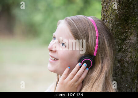 Ragazza seduta contro albero in giardino ascoltando musica sulle cuffie Foto Stock