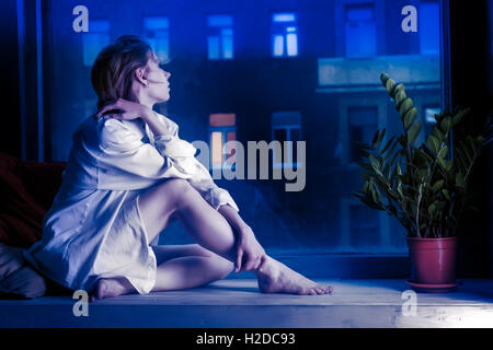 Mezzo vestito ragazza meditativo in maglia bianca si siede sul davanzale. Sera scena urbana al di fuori della finestra. Tonica in colore blu Foto Stock