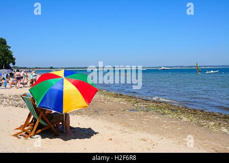 I villeggianti rilassante sulla spiaggia con un colorato ombrellino in primo piano, Studland Bay, Dorset, England, Regno Unito, Europa. Foto Stock