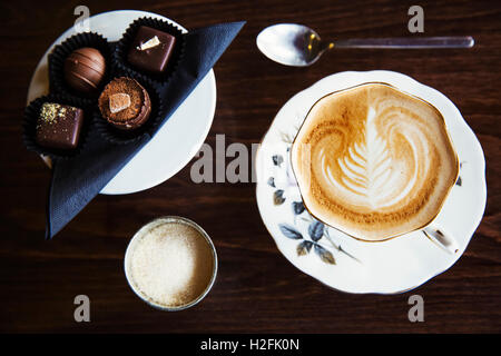 Mano assortiti cioccolatini fatti su una piastra. Una tazza di caffè con un modello in schiuma alto. Foto Stock