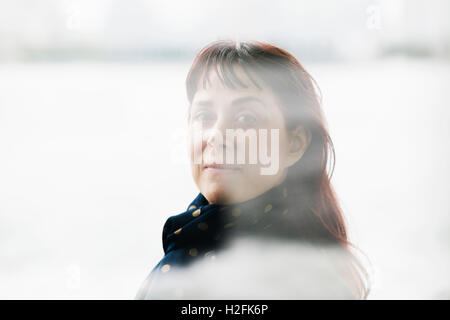 Una donna con capelli castani e una frangia, indossando una sciarpa al suo collo, guardando attraverso una finestra. Foto Stock