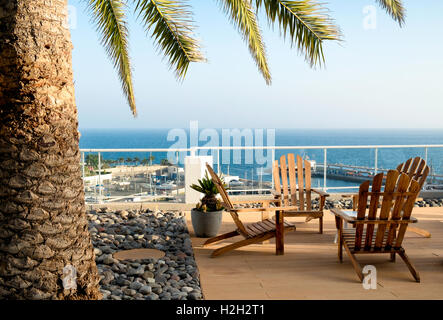Vacation Resort, sedie vuote con l'oceano sullo sfondo e un albero di palma il framing sulla sinistra. Foto Stock