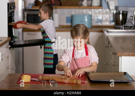 Ragazzo prendendo un vassoio di biscotti al di fuori di un forno mentre una ragazza è il taglio di forme da in prodotti della pasticceria Foto Stock