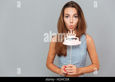 Piuttosto carino giovane donna che soffia sulla torta di compleanno di puntelli su sfondo grigio Foto Stock